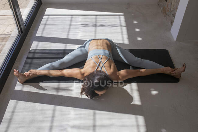Высокий угол анонимной гибкой женщины, исполняющей позу Упависты Конасаны, касаясь ног на коврике для йоги дома — стоковое фото