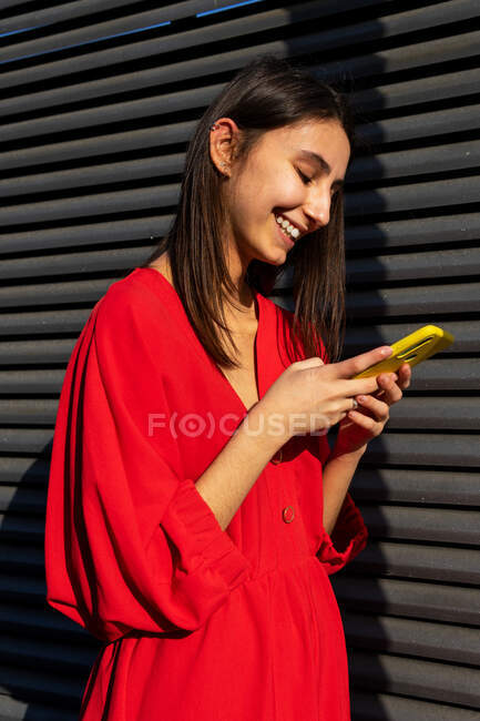 Joven contenido femenino en rojo desgaste chat en el teléfono móvil en la luz del sol sobre fondo gris - foto de stock