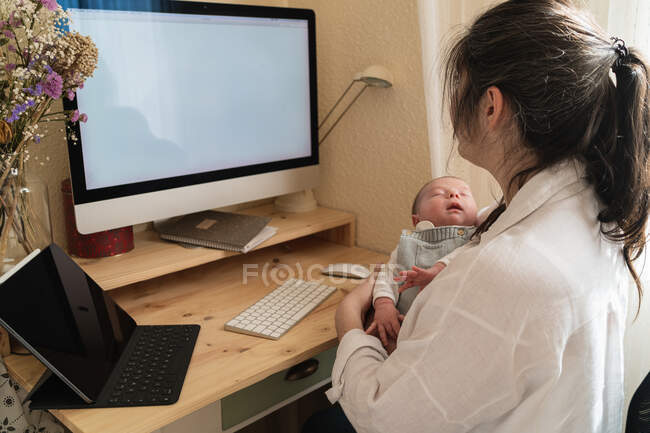 Adult mother sitting at desk working on desktop computer while holding little child at table in daytime — Stock Photo