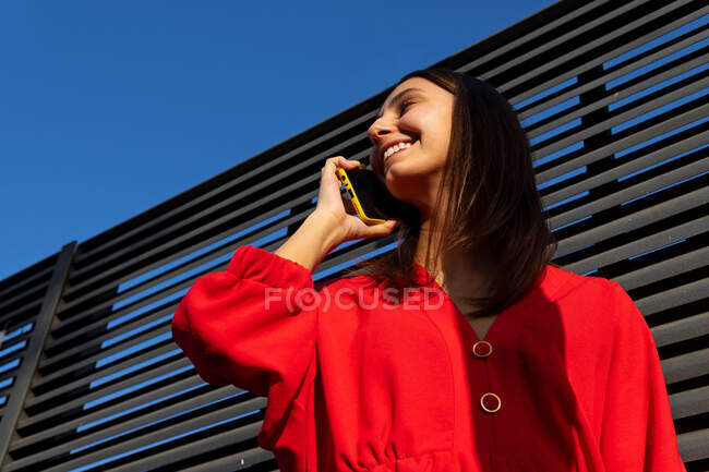 Young content female in red wear talking mobile phone in sunlight on gray background — Stock Photo