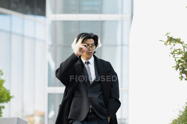 Empresario masculino étnico joven y seguro de sí mismo en traje formal y abrigo caminando con las manos en los bolsillos mientras mira hacia otro lado en la ciudad - foto de stock