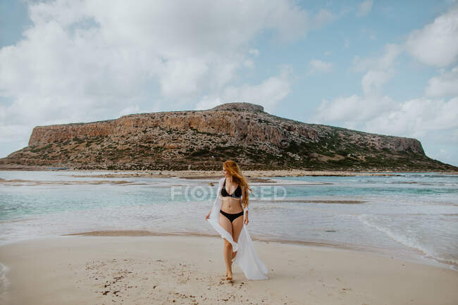 Полная длина стройная женщина в черном купальнике стоит на песчаном пляже Балос против скалистой скалы и смотрит в сторону ясной солнечной погоды — стоковое фото