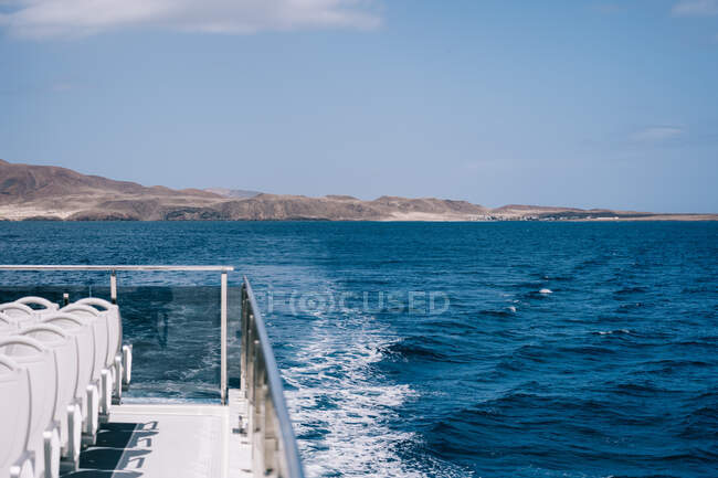 Порожні білі стільці на палубі круїзного човна, що пливе у блакитній морській воді з горою на березі — стокове фото