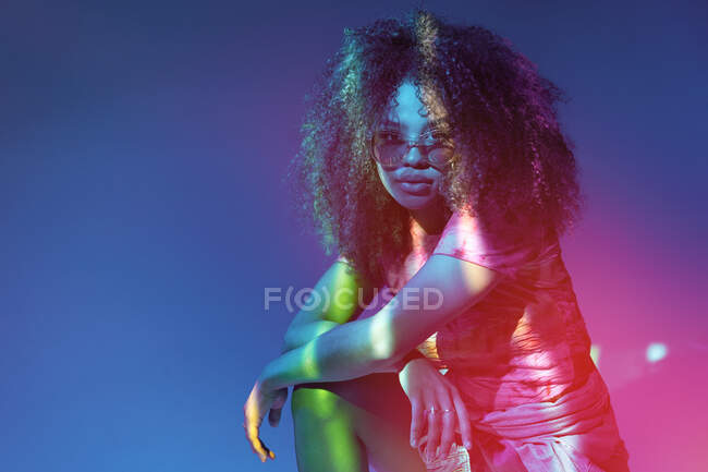 Atractiva joven mujer afroamericana en gafas de sol elegantes mirando a la cámara mientras está de pie en luces de neón - foto de stock