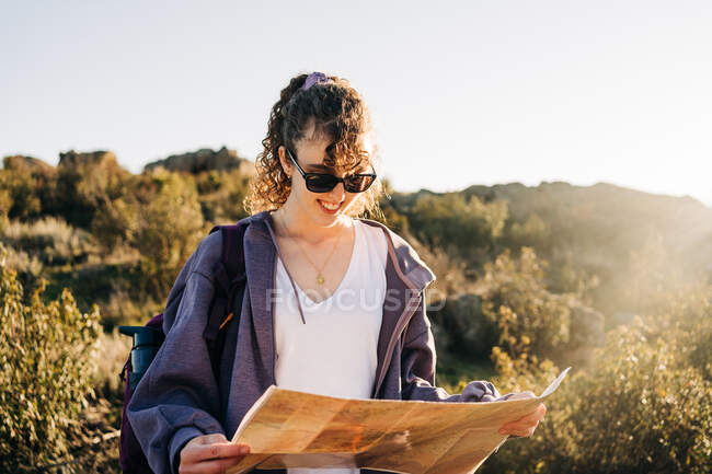 Focused young female explorer in casual clothes reading map while walking on mountain slope on sunny day — Stock Photo