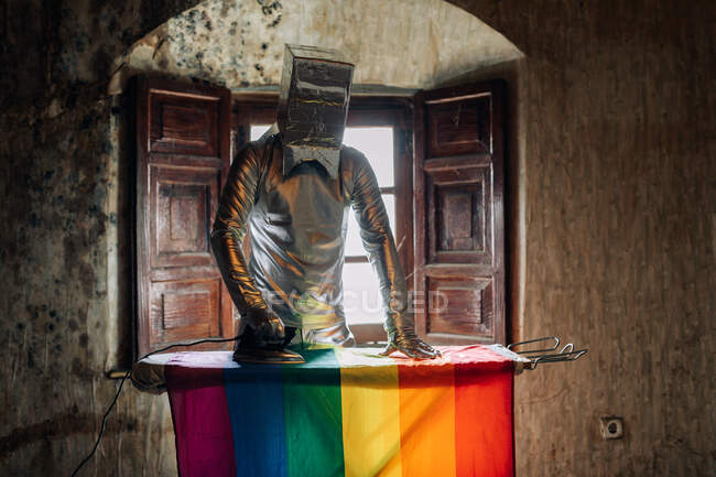 Personne anonyme portant un costume argenté et une boîte sur la tête repassant le drapeau LGBT dans une pièce abîmée de la maison abandonnée — Photo de stock