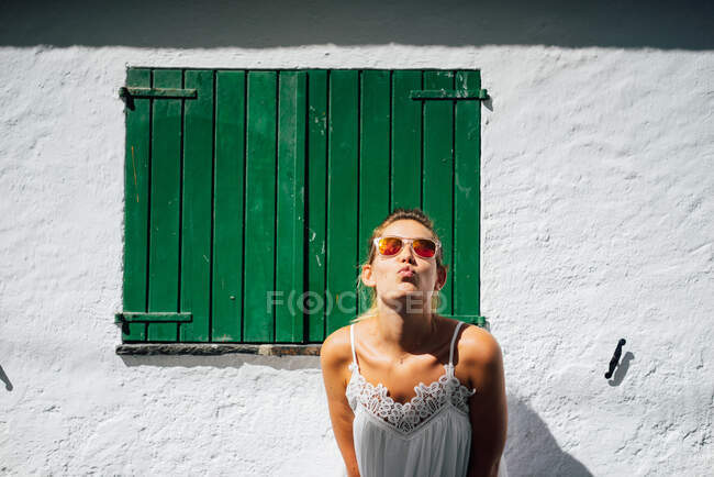 Young female tourist in white apparel and modern sunglasses leaning forward against aged building exterior in sunlight — Stock Photo