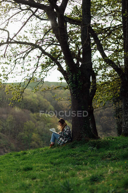 Side view of female traveler with paper guide sitting on meadow between green trees and mount in countryside — Fotografia de Stock