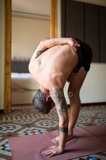 Vista lateral de un hombre sin camisa parado en Ardha Baddha Padmottanasana sobre una esterilla mientras se equilibra y practica yoga en casa - foto de stock