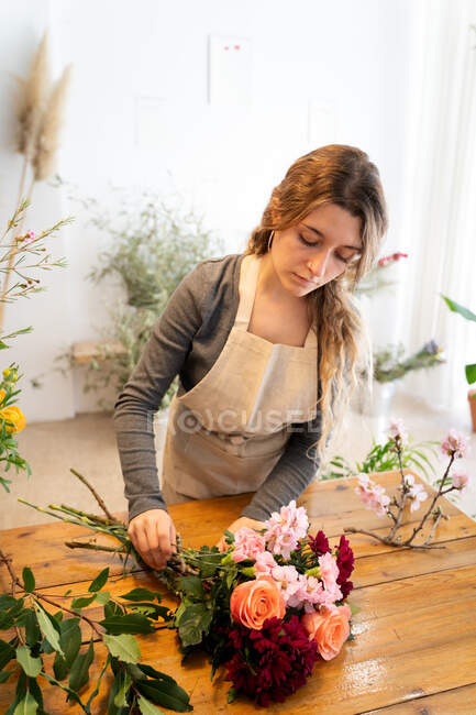 Концентрированная молодая флористка с длинными волнистыми волосами в повседневной одежде и фартуке, раскладывающая элегантный букет роз и разнообразных цветов в магазине — стоковое фото