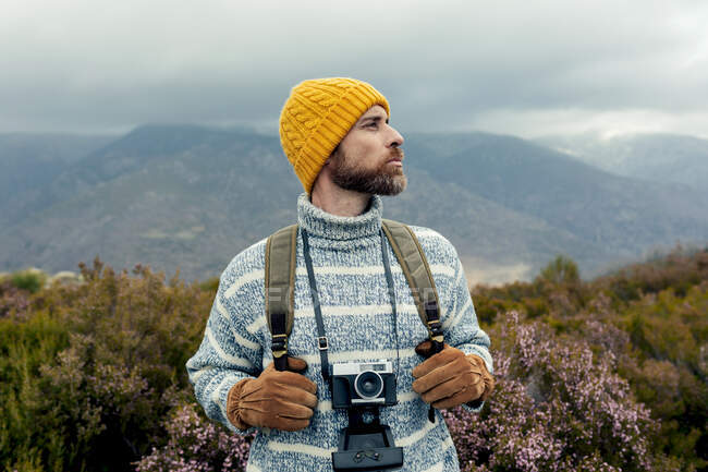 Pensiero camper maschile con cappello caldo in piedi con fotocamera fotografica e zaino in altopiani ammirando e godendo di panorami pittoreschi — Foto stock