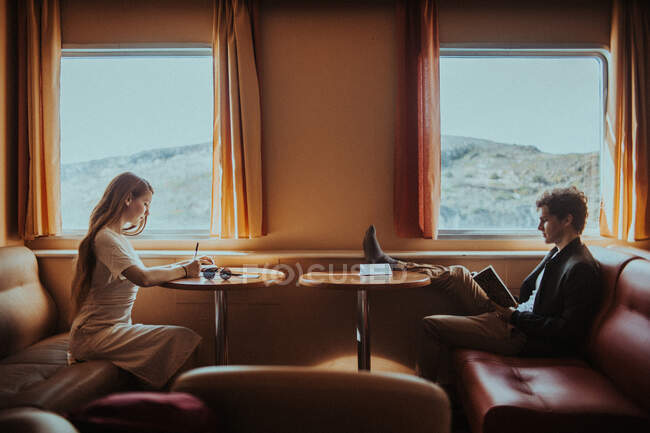 Протилежна молода пара у випадковому літньому одязі читає цікаву книжку і записує думки під час подорожі на поромному човні разом з горбистим узбережжям у сонячній Греції. — стокове фото