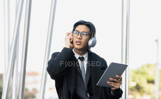 Jeune entrepreneur ethnique masculin dans des écouteurs et des lunettes modernes avec tablette regardant loin en ville — Photo de stock