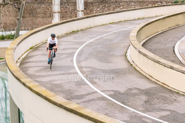 Male bicyclist in sportswear and protective helmet riding bike on curved roadway during training in city — Stock Photo