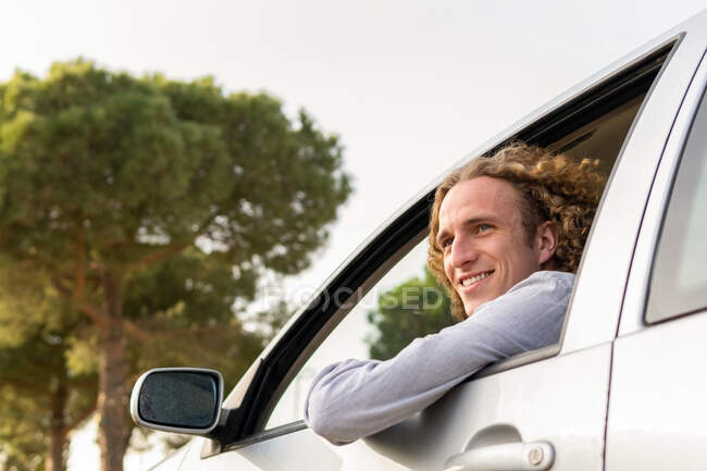 Side view of cheerful young haired male keeping hand out of car window while enjoying summer journey in nature looking away — Photo de stock