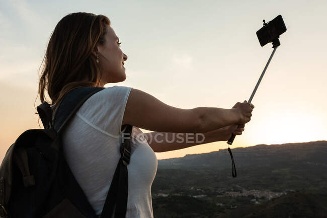 Знизу подорожує самка з рюкзаком, що стоїть на пагорбі і робить самостріл на смартфоні на тлі гірського хребта влітку — стокове фото