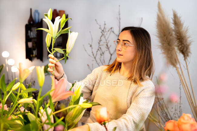 Concentrated young female florist in apron and eyeglasses arranging fragrant flowers in vase while working in floral shop — Stock Photo