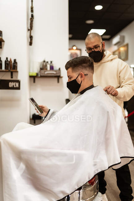 Seitenansicht von Crop Stylist mit Trimmer schneiden Haare des Mannes in Tuch Gesichtsmaske mit Handy im Friseurladen — Stockfoto