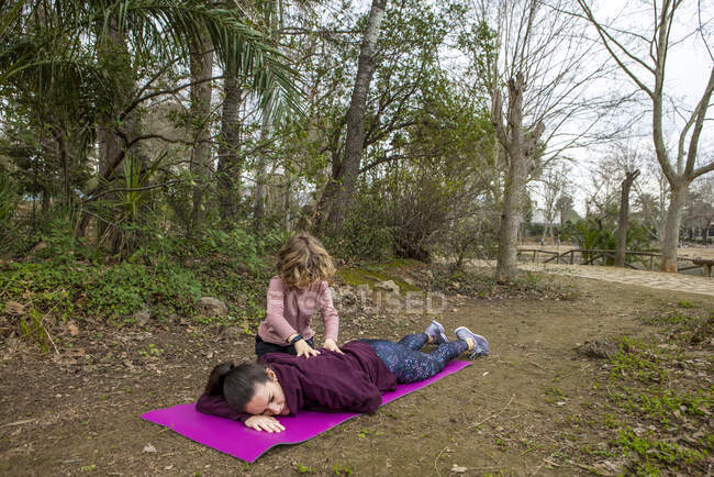 Анонімний дитина масажує спину мами, що лежить на килимку після практики йоги проти дерев у парку — стокове фото