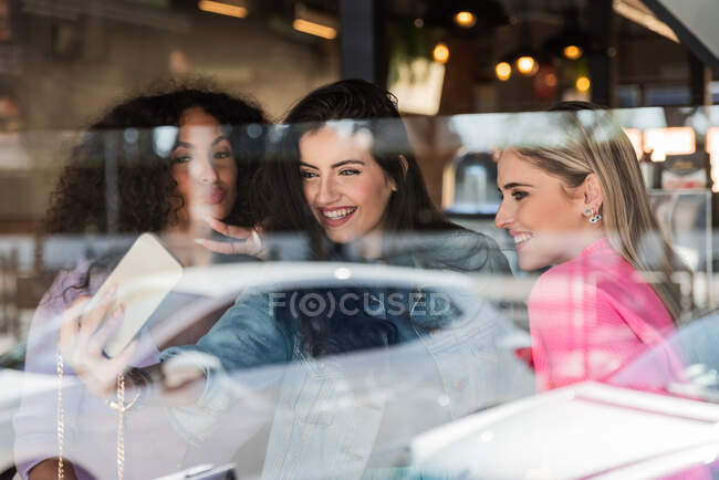 Through glass of cheerful young multiracial female best friends in trendy outfits smiling and showing two fingers gesture while taking selfie on smartphone standing near window — Stock Photo