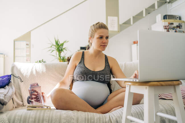 Молода майбутня жінка в спортивному одязі з пляшкою водного серфінгу в Інтернеті на нетбуці, сидячи з схрещеними ногами на дивані — стокове фото