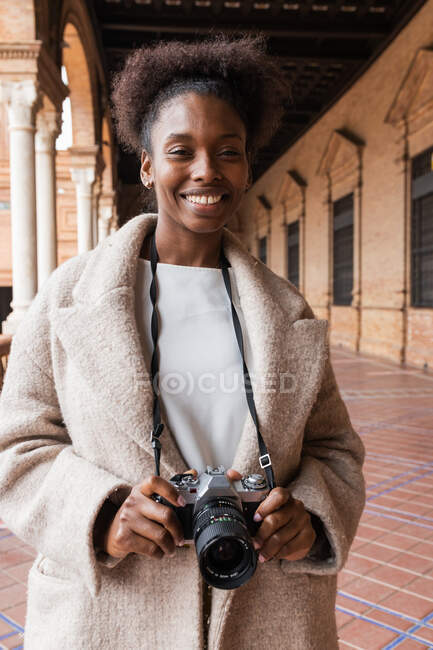 Vista frontal de una joven fotógrafa afroamericana con abrigo cálido en edificios históricos de la ciudad con una cámara fotográfica moderna a principios del día de primavera - foto de stock