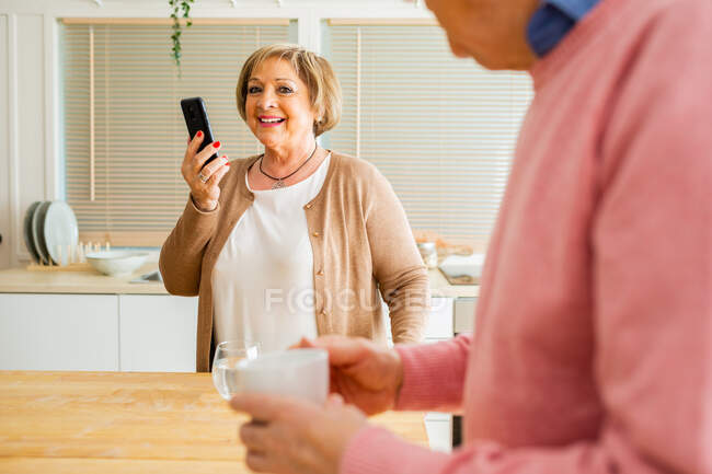 Вміст пари середнього віку, що переглядає планшет і смартфон вранці, стоячи за столом на кухні — стокове фото
