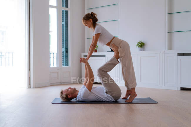 Side view of boyfriend lifting girlfriend while doing acro yoga together at home and holding hands - foto de stock