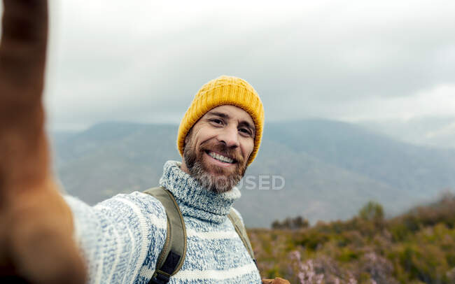 Cheerful bearded male explorer with backpack and in warm clothes taking selfie in mountains and smiling at camera — Stock Photo