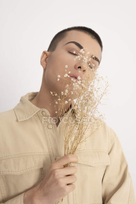 Modello maschio androgeno in camicia alla moda e con mazzo di piante essiccate che guardano la macchina fotografica su sfondo bianco in studio — Foto stock
