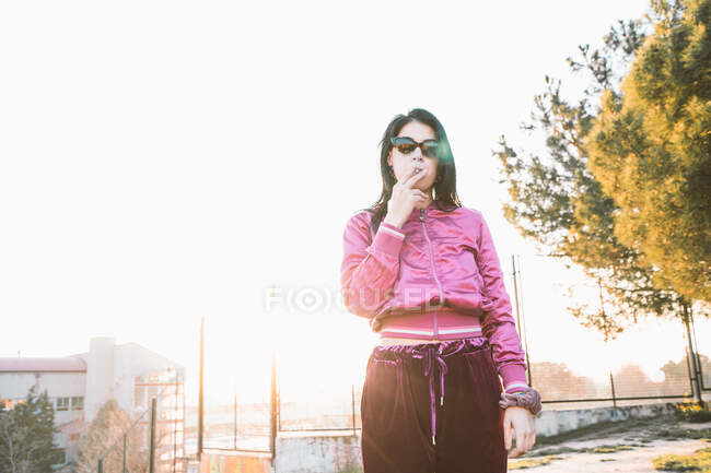 Тисячолітня жінка в модному одязі та сонцезахисних окулярах курить сигарету проти міської будівлі під білим небом — стокове фото