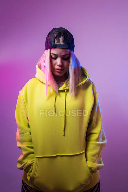 Unemotional female in street style hoodie and cap looking down on purple background in studio with neon illumination — Stock Photo