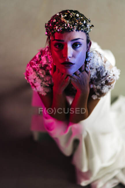 From above of elegant young female in white wedding gown with jewelry wreath looking at camera with praying hands in studio with neon illumination — Fotografia de Stock