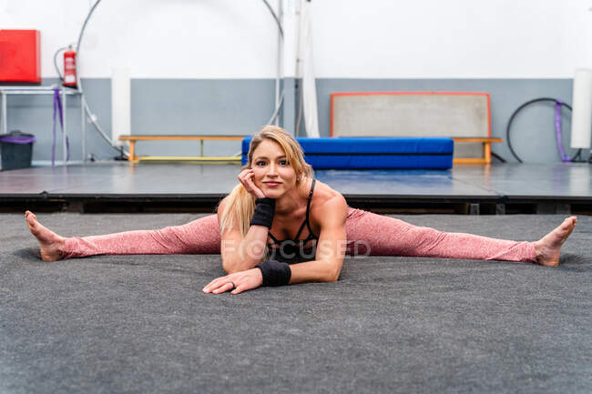 Full body attractive agile female in sportswear doing split and leaning on hand in gymnastic center while looking at camera contentedly — Stock Photo