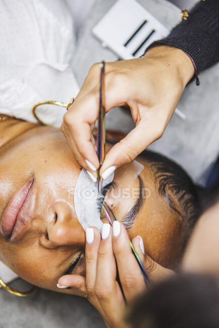 Hoher Winkel der Ernte unkenntlich Kosmetiker mit Pinzette Anwendung gefälschte Wimpern für die Verlängerung auf das Auge des ethnischen Kunden im Salon — Stockfoto