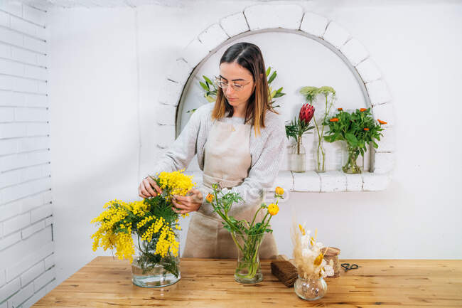 Konzentrierte junge Floristin in Schürze und Brille arrangiert duftende gelbe Mimosenblüten in der Vase, während sie im Blumenladen arbeitet — Stockfoto
