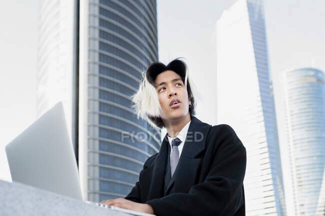 Dal basso giovane dirigente etnico maschio ben vestito con i capelli tinti che lavorano su netbook contro gli edifici moderni della città guardando altrove — Foto stock