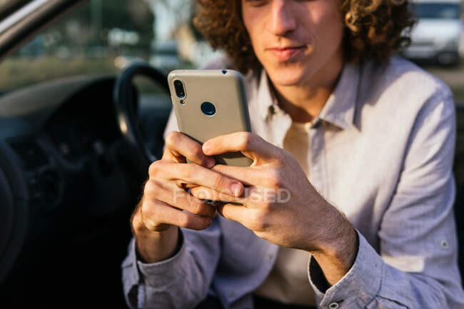 Обрізати молодого чоловіка, сидячи на водійському сидінні сучасного автомобіля з відкритими дверима і повідомленнями на мобільному телефоні, відпочиваючи під час поїздки — стокове фото