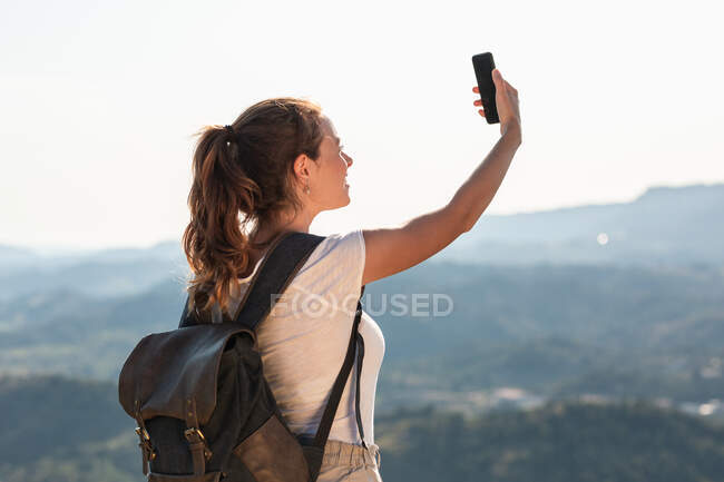 Indietro vista felice giovane escursionista femminile con zaino prendere selfie su smartphone mentre in piedi sulla cima verdeggiante collina — Foto stock