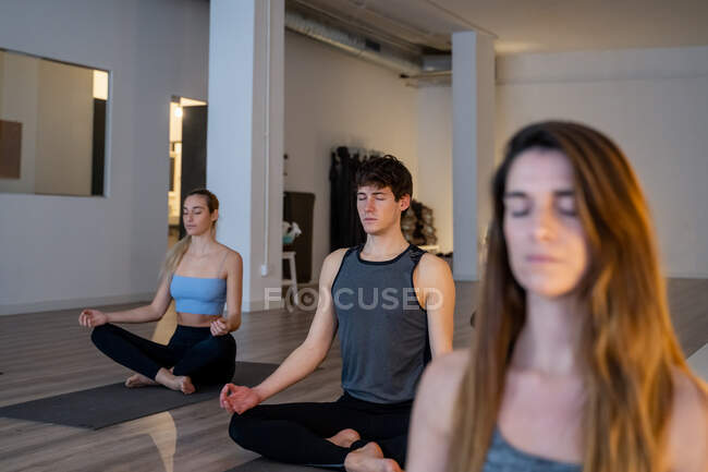 Grupo de diversas personas sentadas en Lotus posan con los ojos cerrados y mediando mientras practican yoga juntos durante la clase en estudio - foto de stock