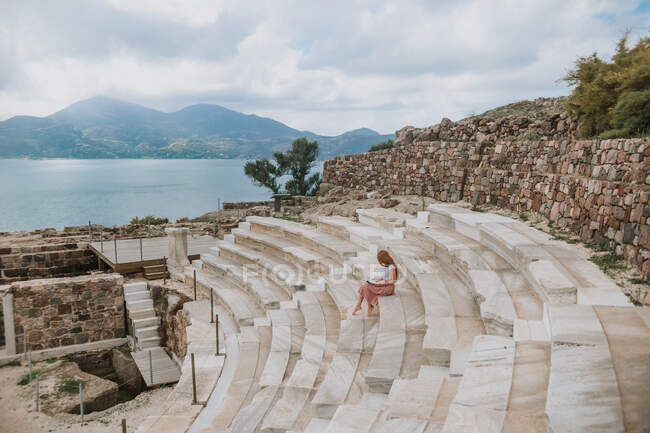 Side view anonymous female in summer wear enjoying good book and sitting on stone steps of ancient theater located on scenic seashore in Milos Greece — Stock Photo