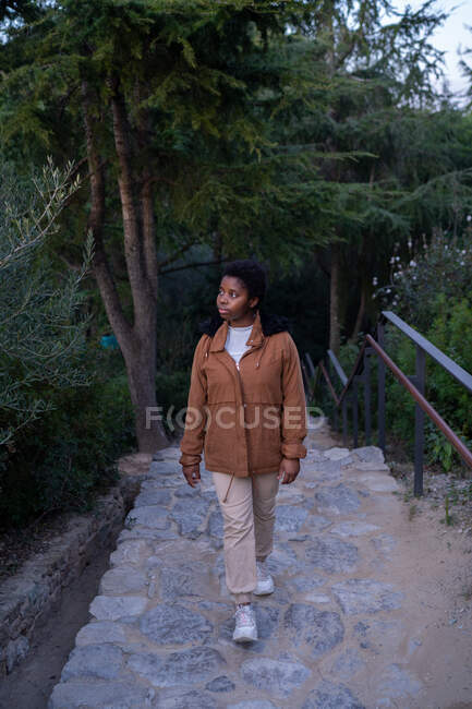 Joven mujer afroamericana tranquila en chaqueta caliente de pie en el verde exuberante parque y mirando hacia otro lado en el día de primavera - foto de stock