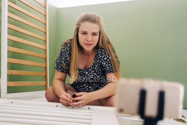 Joven mujer embarazada usando destornillador mientras monta cuna y se sienta en el suelo en casa - foto de stock