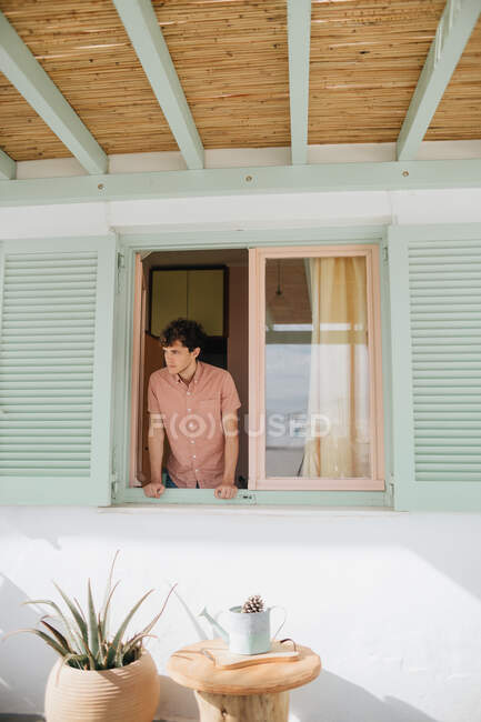 Emotionless jovem macho em camisa casual olhando para fora da janela, enquanto em pé na casa contemporânea em tempo ensolarado — Fotografia de Stock