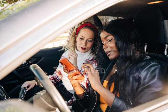 Stilvolle multiethnische Freundinnen sitzen im modernen Auto und surfen in der Stadt auf dem Smartphone — Stockfoto