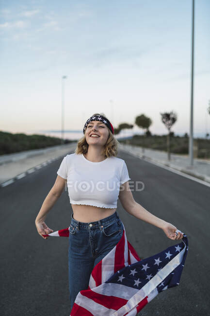 Donna americana deliziata in piedi con bandiera nazionale degli Stati Uniti sulla carreggiata al tramonto e guardando in alto — Foto stock