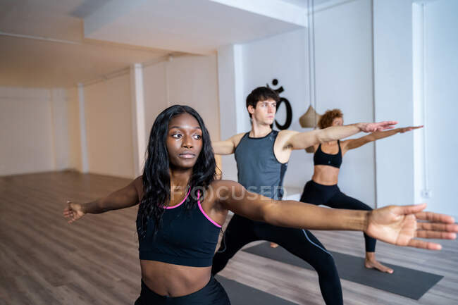 Donna afroamericana in compagnia di diverse persone che praticano yoga in Warrior due posa in studio — Foto stock