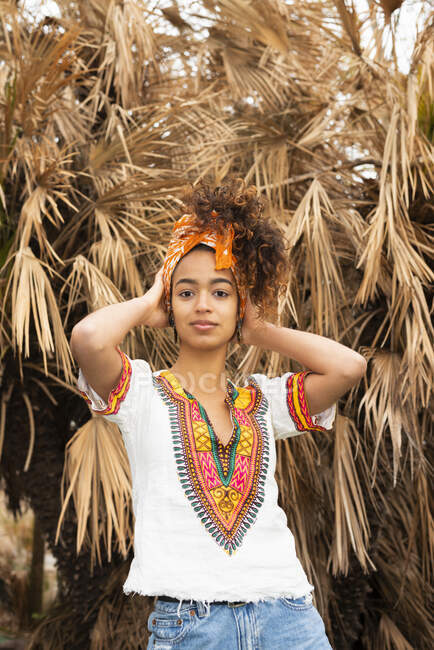 Joven mujer étnica de pie en el prado contra las palmeras y mirando a la cámara - foto de stock