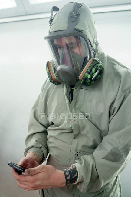 Male worker in respirator and protective uniform standing in car service and browsing mobile phone — Stock Photo