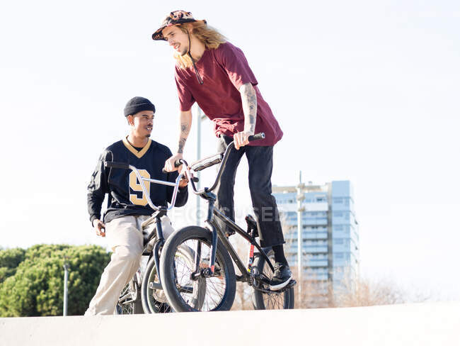 De baixo de jovens desportistas multiétnicos alegres em bicicletas de julgamento olhando um para o outro durante a saudação na cidade — Fotografia de Stock